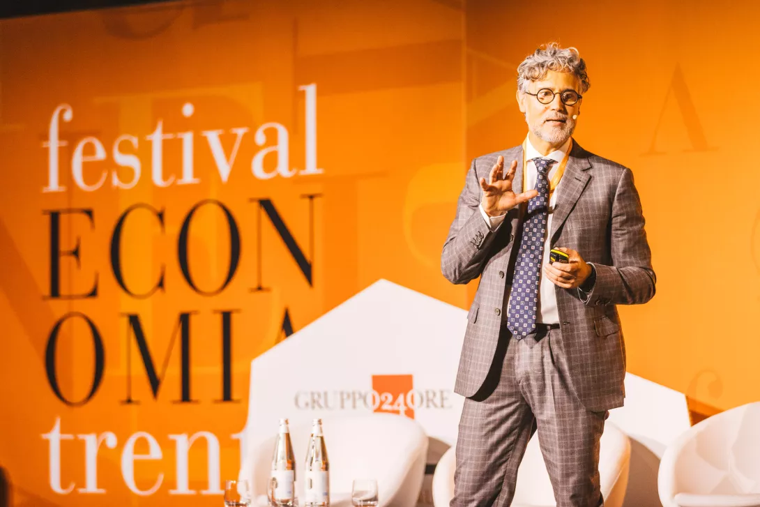 A person presenting on stage at the ‘Festival Economia Trento.’ The individual holds a microphone and gestures while standing in formal attire. The background features large text reading ‘festival economia trento’ and the logo of ‘Gruppo24Ore