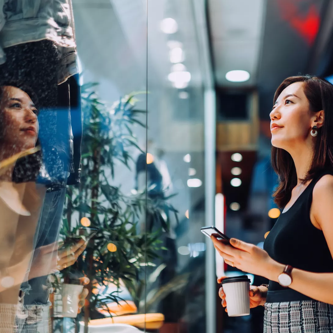 Woman with smartphone and coffee mug in her hands looking at a shop window